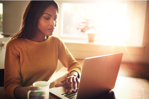 A woman in a yellow top typing on a laptop at a desk, holding a cup of coffee, with sunlight streaming through the window